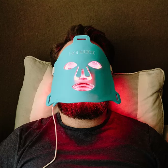 A person with a beard lies on a pillow wearing a blue LED light therapy face mask that glows red in several areas, connected to a power source by a white cord.