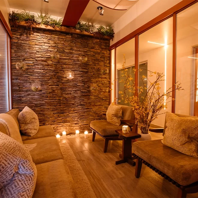 A cozy spa lounge with soft lighting, plush beige seating, decorative pillows, a stone accent wall, candles, and yellow flowers, creating a warm and relaxing atmosphere.