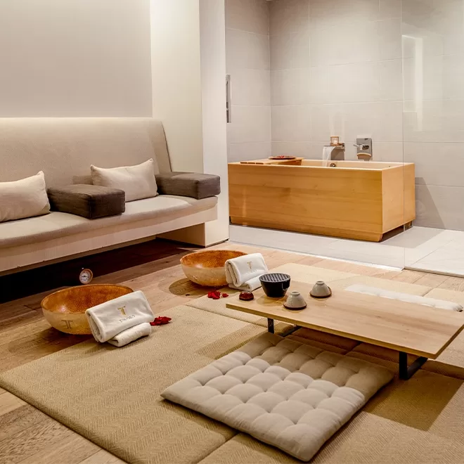 A serene spa room with cushioned floor seating, a low wooden table set with tea cups, a beige sofa with gray pillows, wooden foot soak bowls with towels, and a wooden soaking tub in the background.