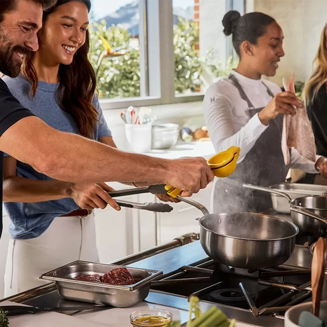 Three people cook together in a bright kitchen. A man squeezes a lemon over a pot as a woman beside him smiles. Another woman in the background prepares ingredients at the counter.