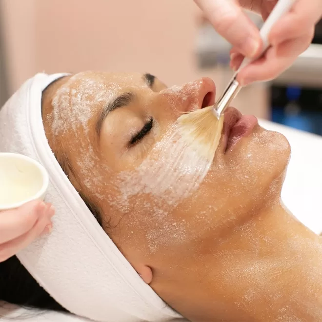 A person with a white headband is lying down while a facial treatment is applied to their face with a brush by another person. Their eyes are closed and their face is covered in a creamy mask.