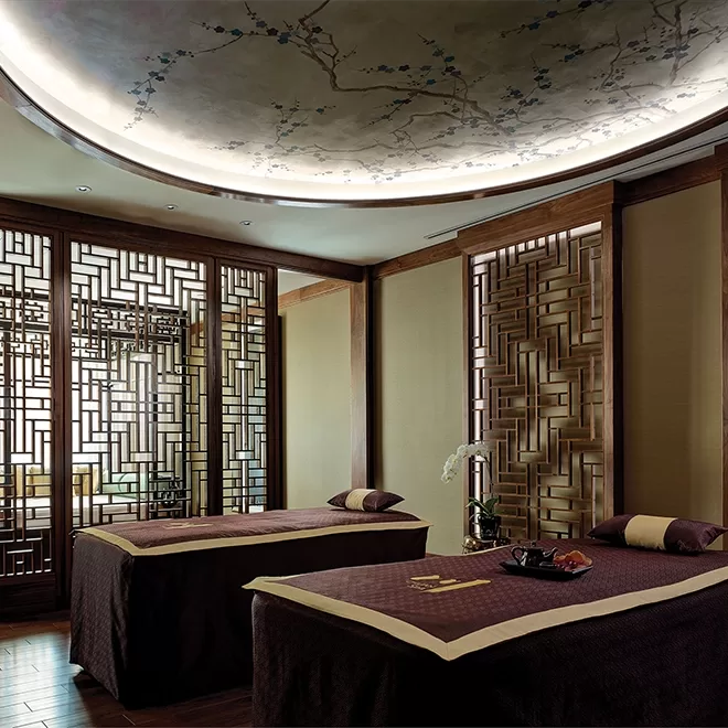 A serene spa room with two massage tables, decorative wooden screens, soft lighting, and a ceiling painted with delicate floral branches, creating a calming and elegant atmosphere.