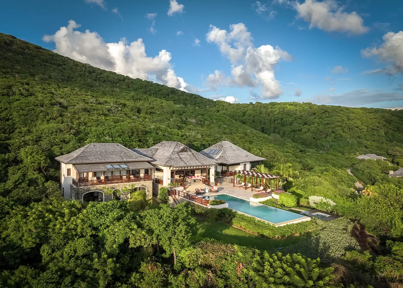 A large luxury villa with a pool sits surrounded by lush green hills under a blue sky with scattered clouds. The house features multiple roofs and balconies, with landscaped gardens and outdoor seating areas.