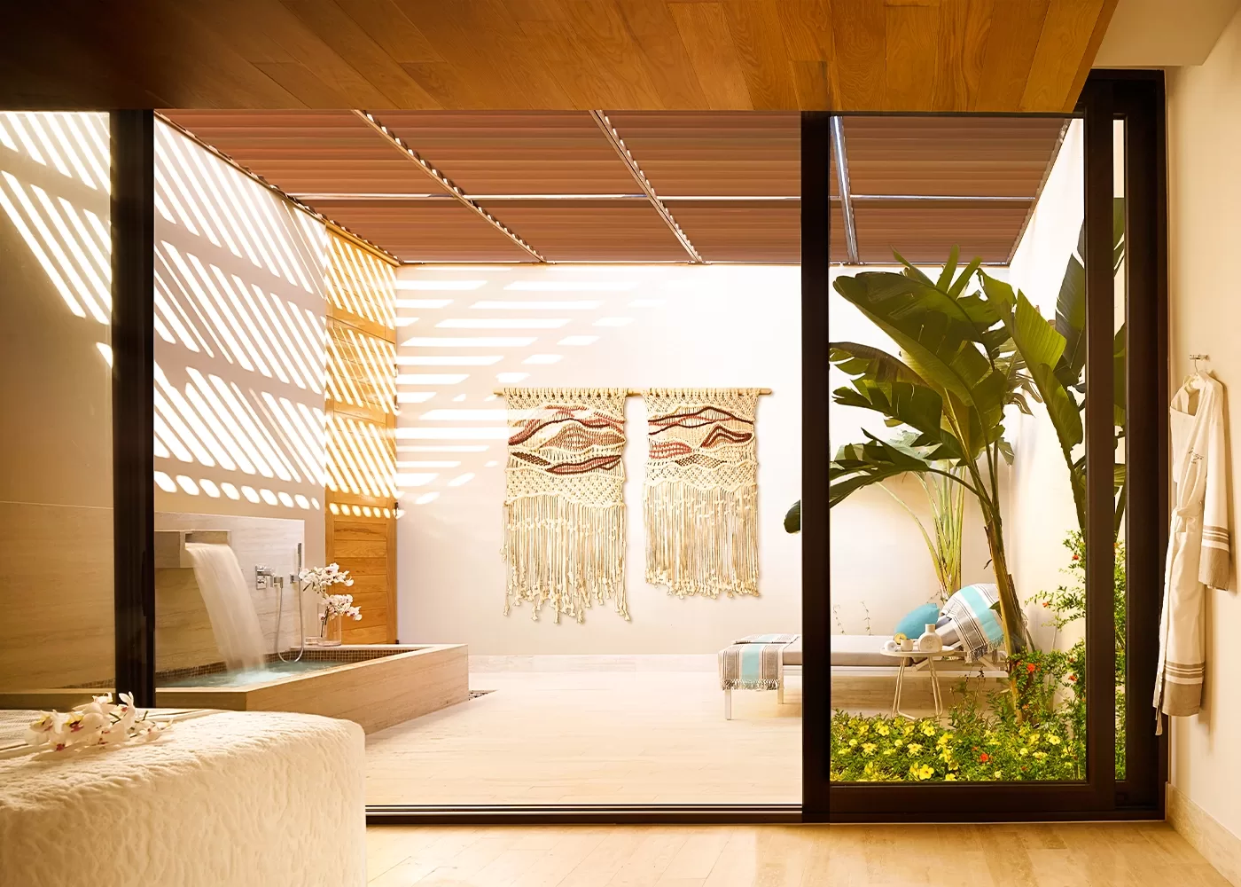 Modern indoor-outdoor bathroom with a soaking tub, waterfall faucet, woven wall hangings, tropical plants, a lounge chair, and sunlight streaming through slatted wooden panels onto light wood floors.