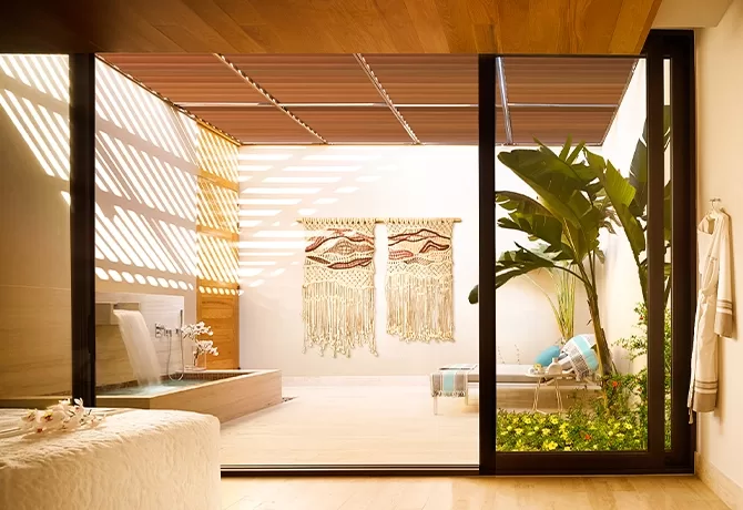 A sunlit indoor space features a lounge chair, tropical plants, a small water fountain, and two decorative wall hangings. Natural light streams through slatted wooden panels, creating shadows on the floor and walls.