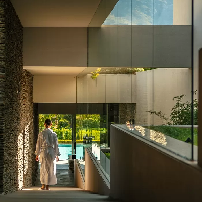 A person in a white robe walks down a sunlit corridor with stone and glass walls, heading toward a lush garden and a blue swimming pool visible through large windows.