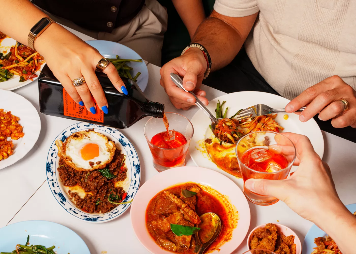 Three people share a colorful meal at a table, serving dishes like curry, a fried egg over minced meat, and salad. One person pours a dark drink into a glass while others toast or eat with forks.