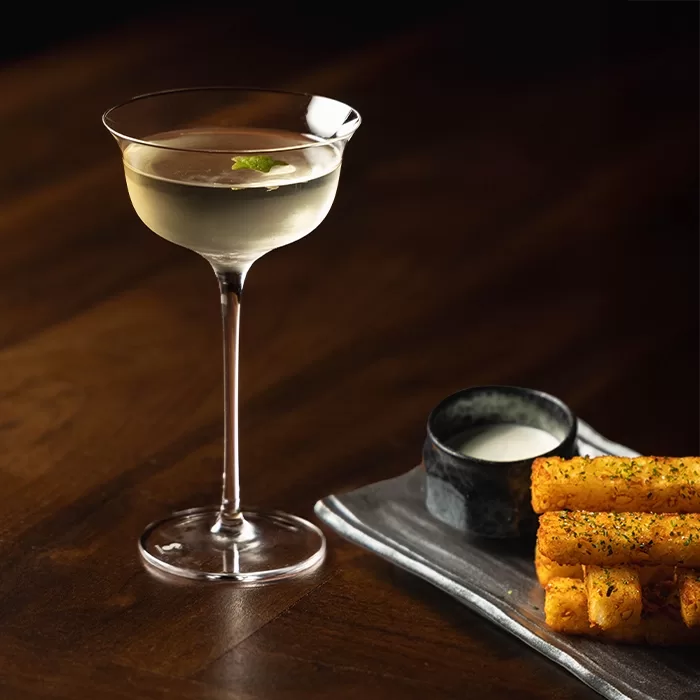 A clear cocktail garnished with a mint leaf in a coupe glass sits next to a rectangular plate with golden, seasoned fries and a small bowl of dipping sauce on a dark wooden surface.