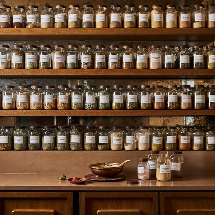 Clear glass jars filled with various spices and herbs are neatly arranged on wooden shelves above a kitchen counter, which holds a copper pan, three jars, and spoons with spices.