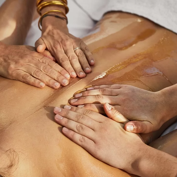 Three pairs of hands gently massaging oil onto a persons bare back, suggesting a relaxing spa or wellness treatment. The person receiving the massage is lying down with a white towel nearby.