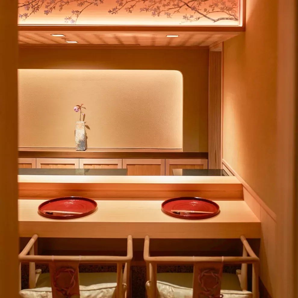 A cozy dining area with two wooden chairs facing a counter adorned with two red plates. Soft lighting highlights a minimalist setting, with a single decorative vase holding a flower, and a floral design on the ceiling panel.