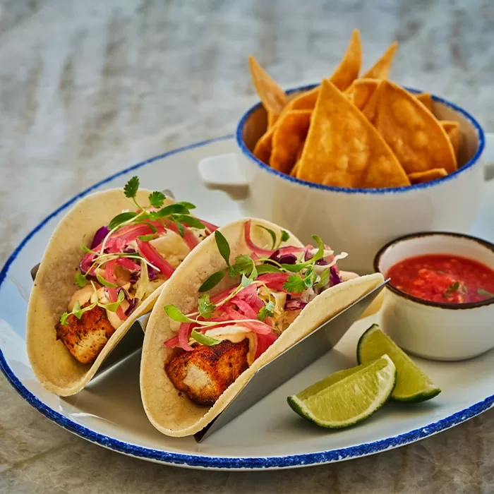 Two fish tacos topped with slaw and pickled onions on a plate, served with lime wedges, a bowl of tortilla chips, and a small dish of red salsa.