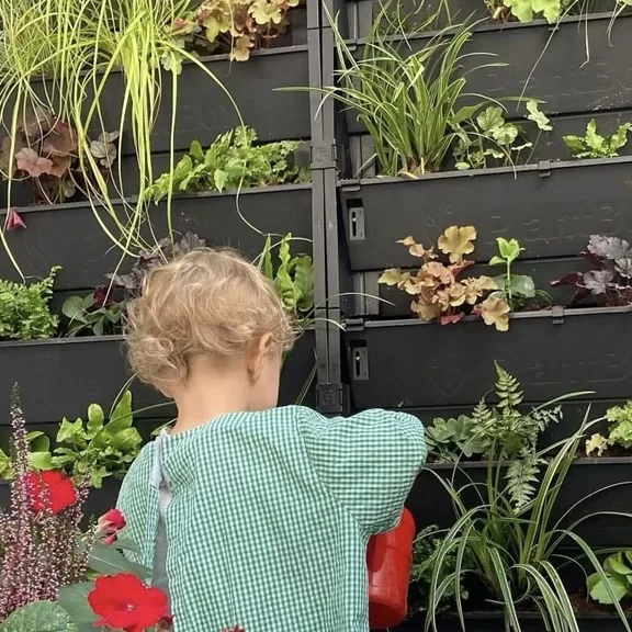 A young child with curly blonde hair and a green checked shirt waters plants in front of a vertical garden filled with various green and leafy plants.