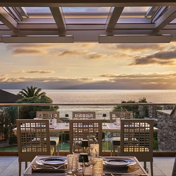 Outdoor restaurant table set for dinner, overlooking a scenic ocean view with a sunset sky, palm tree, and lush greenery beyond a glass railing.