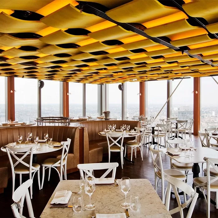 The modern restaurant interior of Duck & Waffle, Bishopsgate in London - with large windows offering a city view. It features a distinctive yellow and black ceiling design, white chairs, wooden tables with place settings, and cosy brown booth seating.