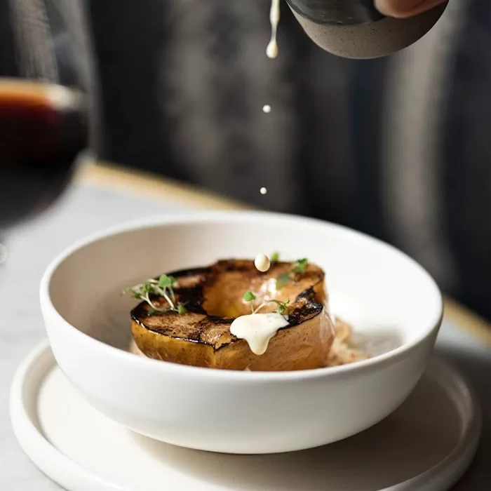 A halved, roasted squash in a white bowl is garnished with fresh herbs and cream. Cream is being poured over the top, with droplets captured in motion. The dish is set on a white plate against a softly blurred background.