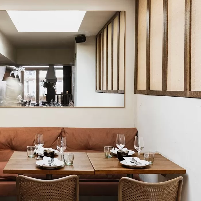 Cozy restaurant interior with a soft brown bench, two wooden tables set with glasses and plates, and wicker chairs. A large mirror on the wall reflects the ambient light, enhancing the warm, inviting atmosphere.