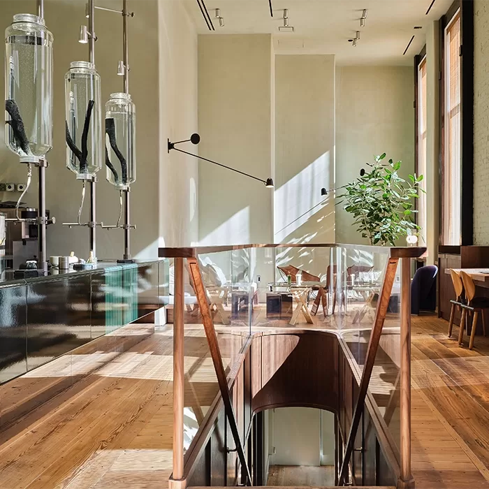 A modern café interior with wood floors, large windows, a glass railing around stairs, and a minimalist counter featuring tall glass brewing equipment. Sunlight streams in, illuminating tables, chairs, and a green potted plant.