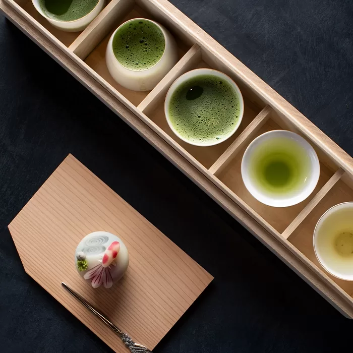 A wooden tray holds five cups of green tea in varying shades, next to a wooden plate with a decorative Japanese wagashi sweet and a fork, all set on a dark surface.
