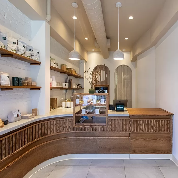 A minimalist café interior with wooden counters, white walls, shelves holding coffee and tea products, pendant lights, and a small display case on the counter. Tall vases and plants add a touch of greenery.
