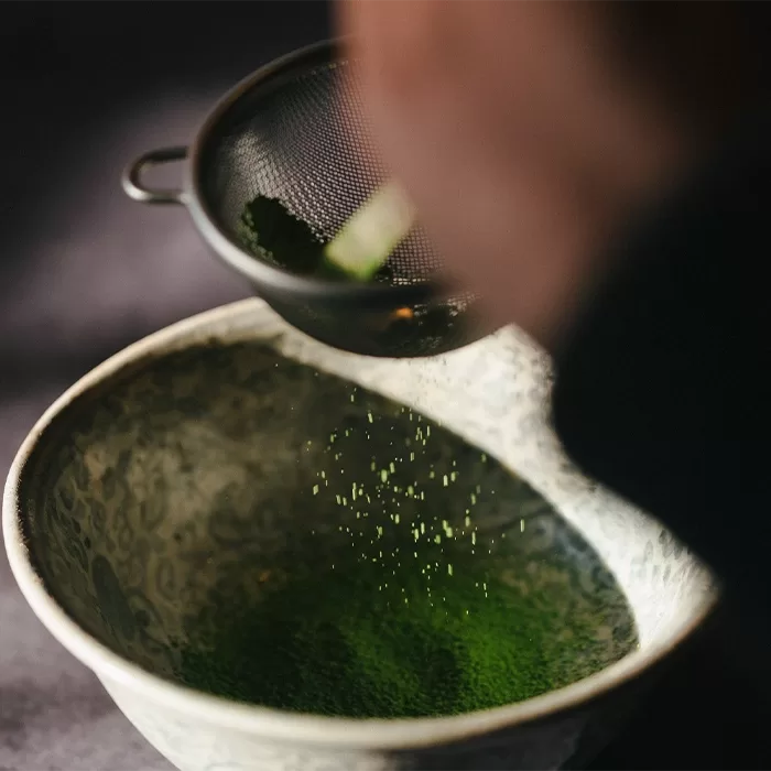 A close-up of a hand sifting vibrant green matcha powder through a fine mesh strainer into a patterned ceramic bowl.