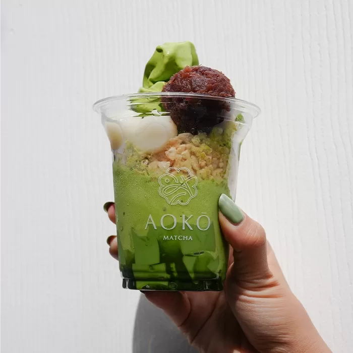 A hand holds a clear cup filled with green matcha dessert topped with whipped cream, a scoop of red bean paste, crispy cereal, and a swirl of matcha soft serve against a white background. The cup reads AOKO MATCHA.