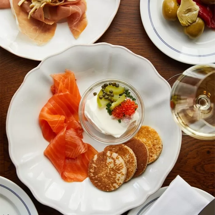A plate with smoked salmon slices, small pancakes, and a glass dish of sour cream topped with capers and fish roe, next to a glass of white wine and other appetizer plates on a wooden table.