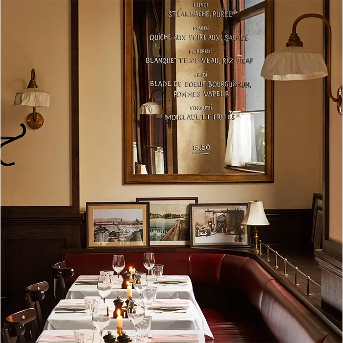 A cozy restaurant interior with a set dining table, white tablecloth, lit candles, and wine glasses. Framed photos are displayed against the wall beneath a large mirror with text listing menu items in French.