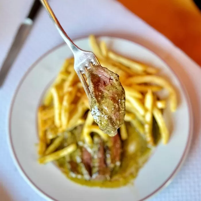A fork holds a piece of steak covered in green sauce above a plate of French fries and steak with green sauce on a white tablecloth.