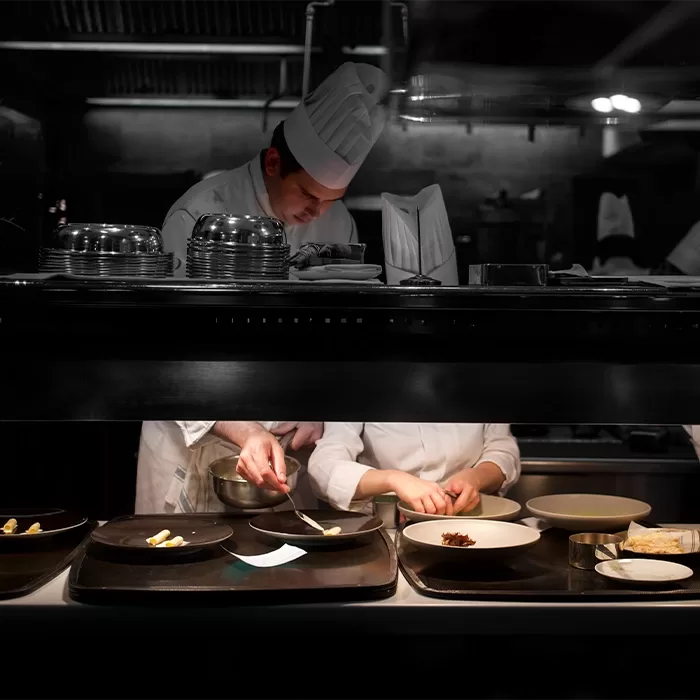 Two chefs wearing white uniforms and tall hats work in a dimly lit kitchen, carefully plating food on black dishes under a bright overhead light. Various kitchen tools and food items are visible on the counter.