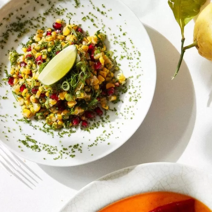 A white plate with a colorful salad made of corn, pomegranate seeds, greens, and herbs, garnished with a wedge of lime and sprinkled herbs, set on a white table with part of a fork and another plate visible.