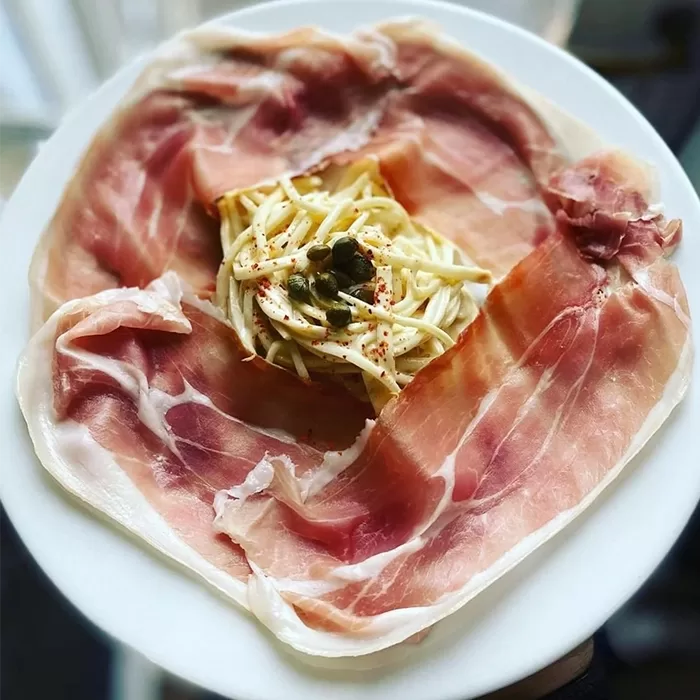 A white plate with a serving of thinly sliced prosciutto arranged in a circle around a small nest of spaghetti topped with capers and seasoning.