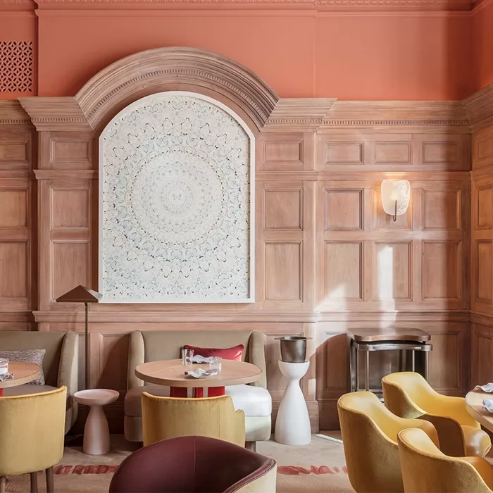 A sophisticated lounge with salmon-colored walls, wood paneling, modern yellow and beige chairs, round tables, and a large intricate white artwork framed by an arched molding above the seating area.