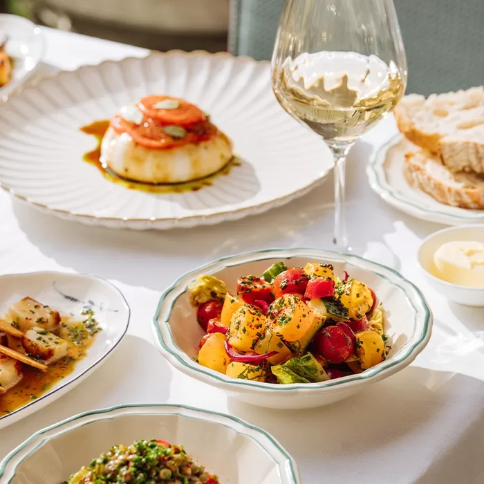 A table set with various dishes including a bowl of colorful tomato salad, a glass of white wine, bread slices, butter, and other plated appetizers on a white tablecloth.