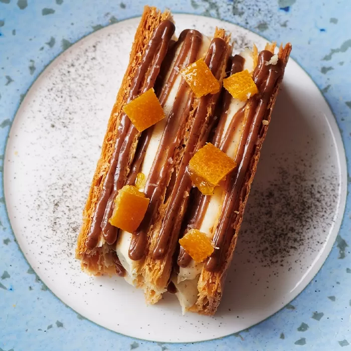 A dessert with layers of flaky pastry, cream, and chocolate drizzle, topped with small orange candied fruit pieces, is served on a white plate with a blue speckled rim and a dusting of dark powder.