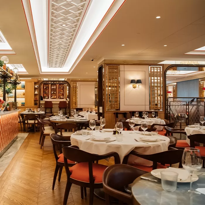 Elegant restaurant interior with round tables set with white tablecloths, wine glasses, and wooden chairs. Warm lighting and decorative ceiling panels create a cozy, upscale atmosphere. The space is empty of people.