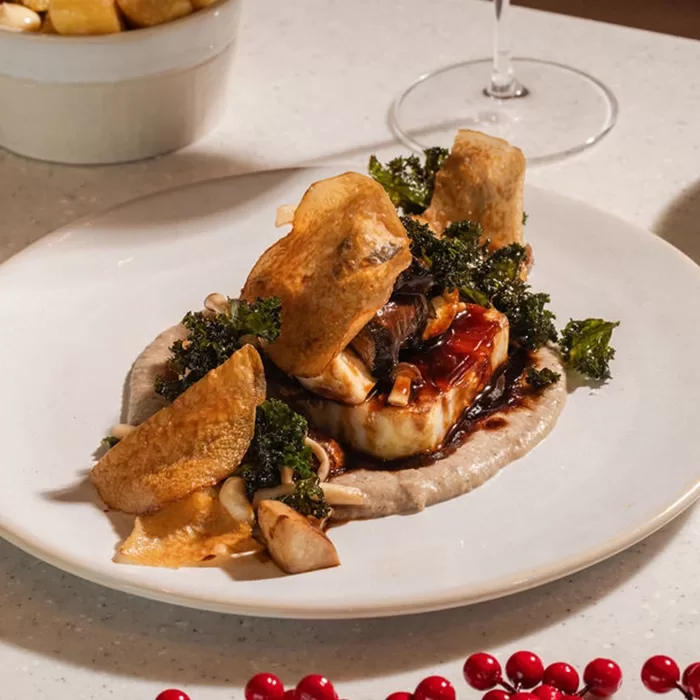 A gourmet dish featuring a piece of fish on a creamy purée, garnished with crispy chips, sautéed mushrooms, and kale, drizzled with sauce. A glass of wine and a bowl of fries are in the background.