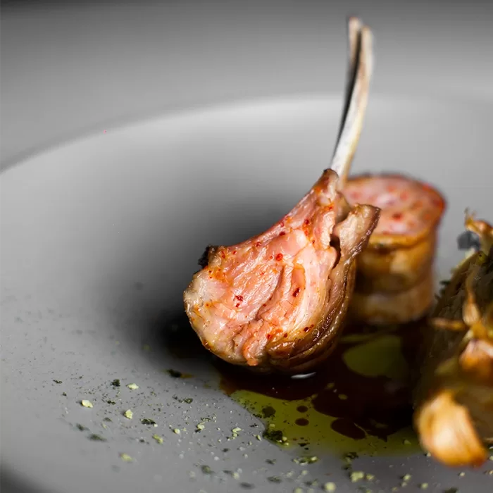 A close-up of a cooked lamb chop, juicy and pink inside, resting on a white plate with a drizzle of sauce and a sprinkle of herbs. Another piece of lamb is visible in the background.