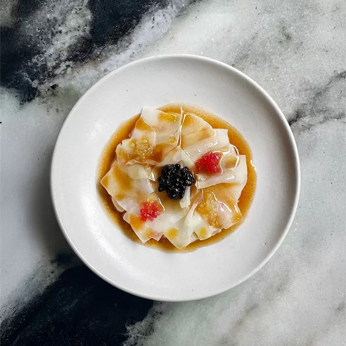 A white plate with thinly sliced raw fish arranged in a circle, topped with caviar and small dollops of red garnish, all drizzled with a light brown sauce, set on a marble surface.