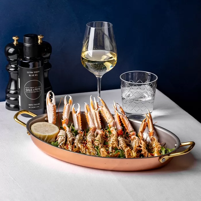 A copper dish filled with cooked langoustines garnished with herbs and a lemon wedge sits on a white tablecloth beside a glass of white wine, a glass of water, black pepper grinder, and an olive oil bottle.