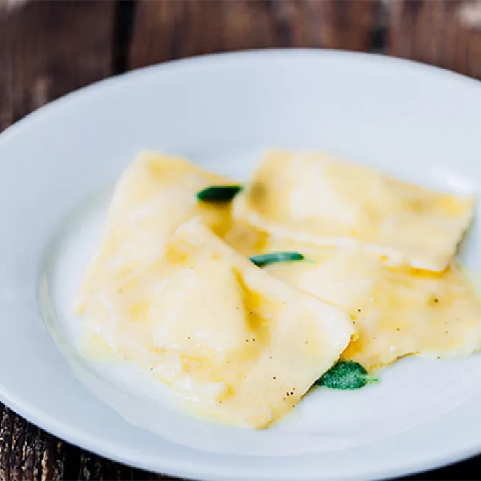 Three pieces of ravioli topped with a light butter sauce and garnished with fresh sage leaves, served on a white plate over a rustic wooden table.