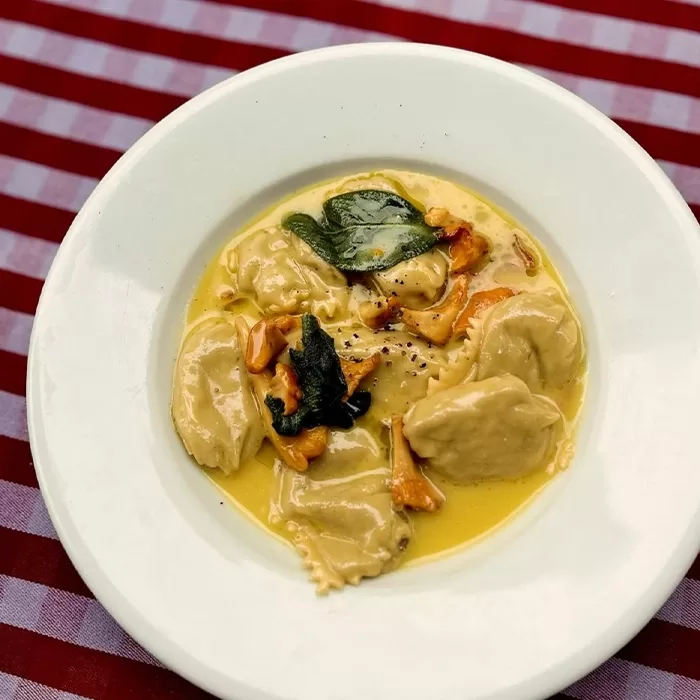 A white plate with four ravioli in a creamy yellow sauce, garnished with fresh sage leaves and cracked black pepper, on a red and white striped tablecloth.