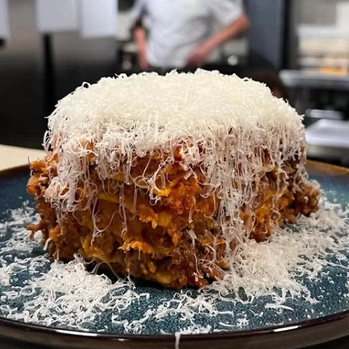 A thick slice of lasagna topped with a large amount of finely grated cheese sits on a blue plate; a chef stands blurred in the background of a kitchen.