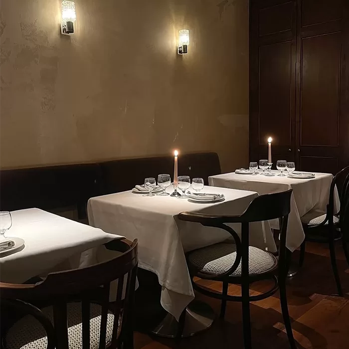 Three tables set for dinner in a dimly lit restaurant, each with a white tablecloth, neatly arranged glassware, and a single lit candle. Dark wooden chairs surround the tables, creating an intimate atmosphere.