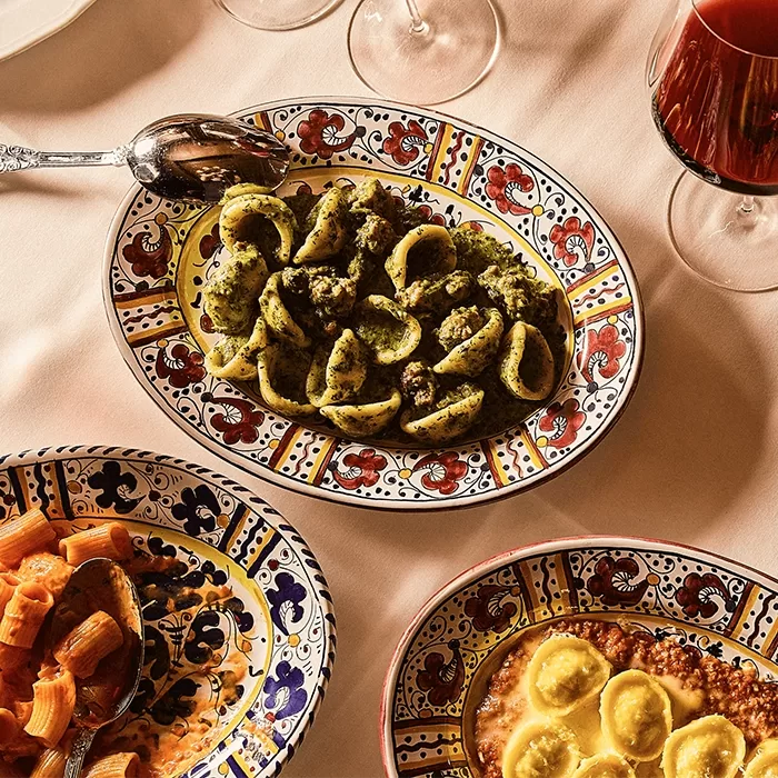 Colorful plates of Italian food, including orecchiette pasta with pesto, ravioli in sauce, and rigatoni, are arranged on a table with wine glasses, all set on a beige tablecloth.
