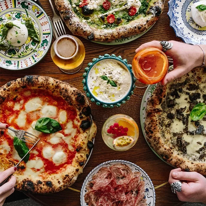 Four pizzas with different toppings, a plate of sliced cured meat, a dish of dip, a salad, and various drinks on a wooden table. Two hands are reaching in, one cutting pizza and the other holding a glass.