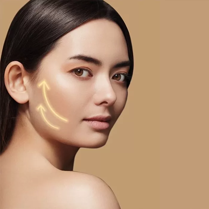 A woman with smooth skin looks over her shoulder against a beige background. Two glowing yellow arrows are drawn on her cheek, pointing upward toward her ear, suggesting a skin tightening or lifting effect.