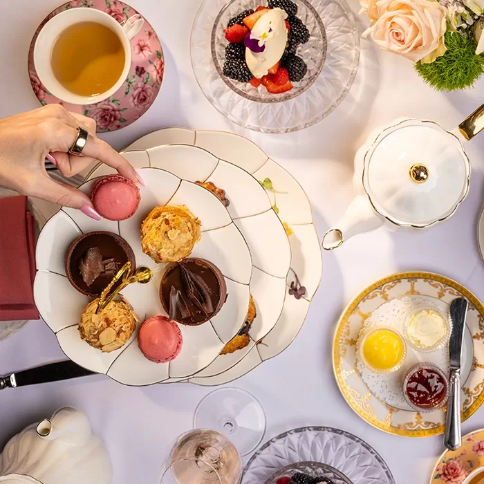 An elegant afternoon tea setup with a hand picking a pink macaron from a tiered tray filled with assorted pastries, surrounded by teapots, floral china, cream, jam, and fruit-topped desserts on a white tablecloth.
