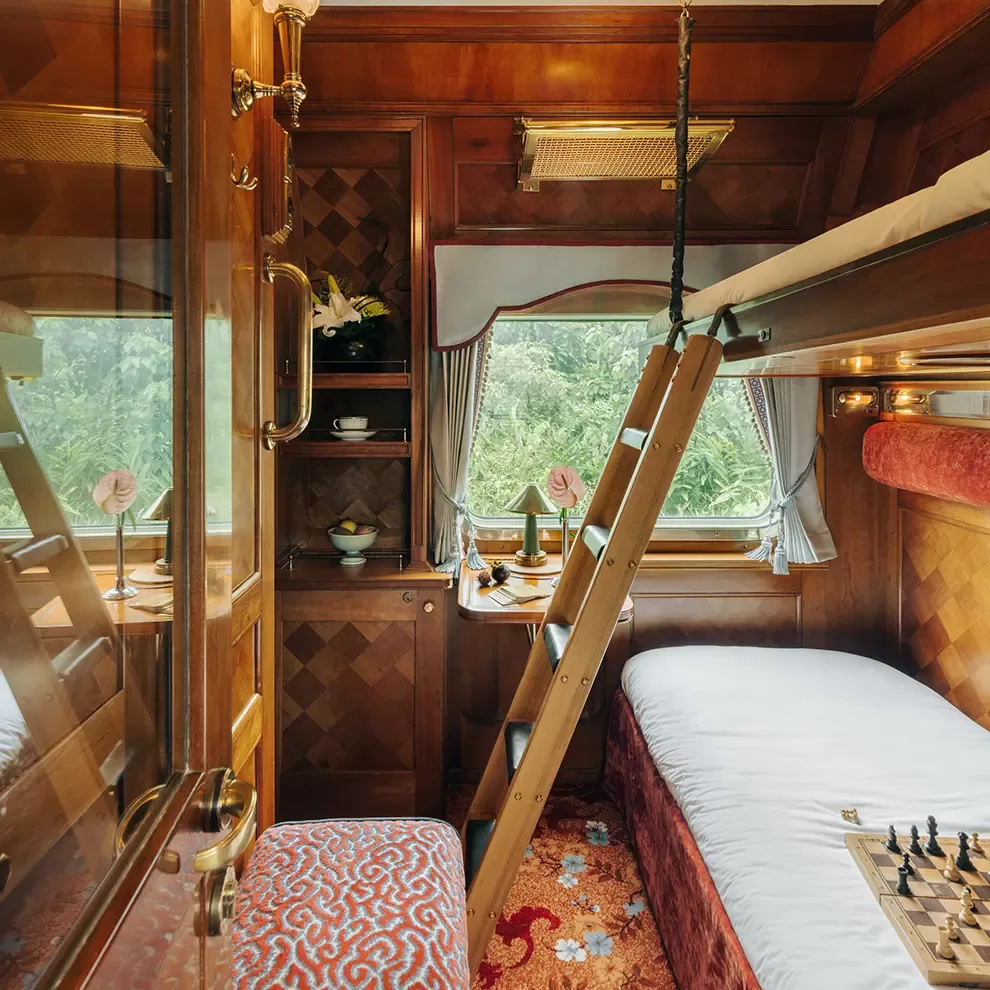 Luxurious train cabin of Eastern & Oriental Express, A Belmond Train in Southeast Asia featuring polished wood panels, a bed with a white sheet, an overhead bunk with a wooden ladder, and large window with forest views. A small table holds a chessboard, dishes, and curtains with tassels adorn the window.