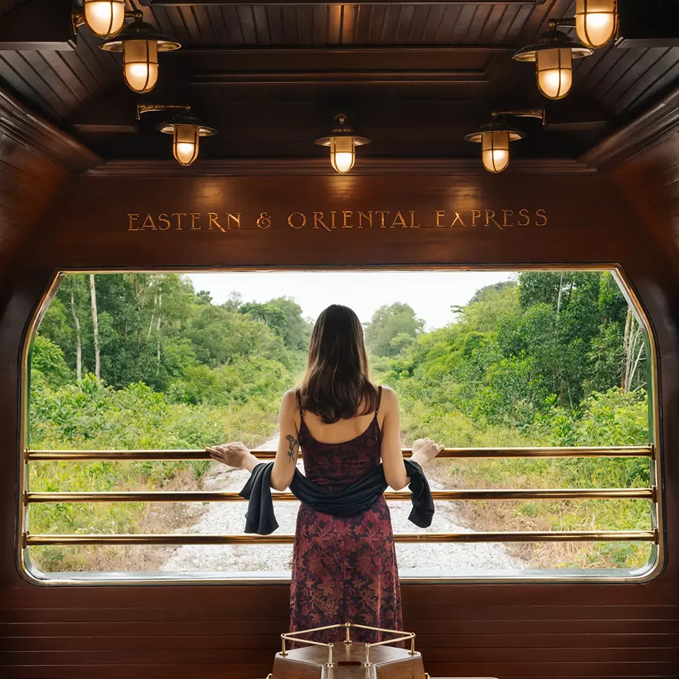 A woman stands at the open back of the Eastern & Oriental Express train, gazing at lush green jungle scenery. She is wearing a patterned dress and holds a jacket. The train interior is wooden with warm lighting.